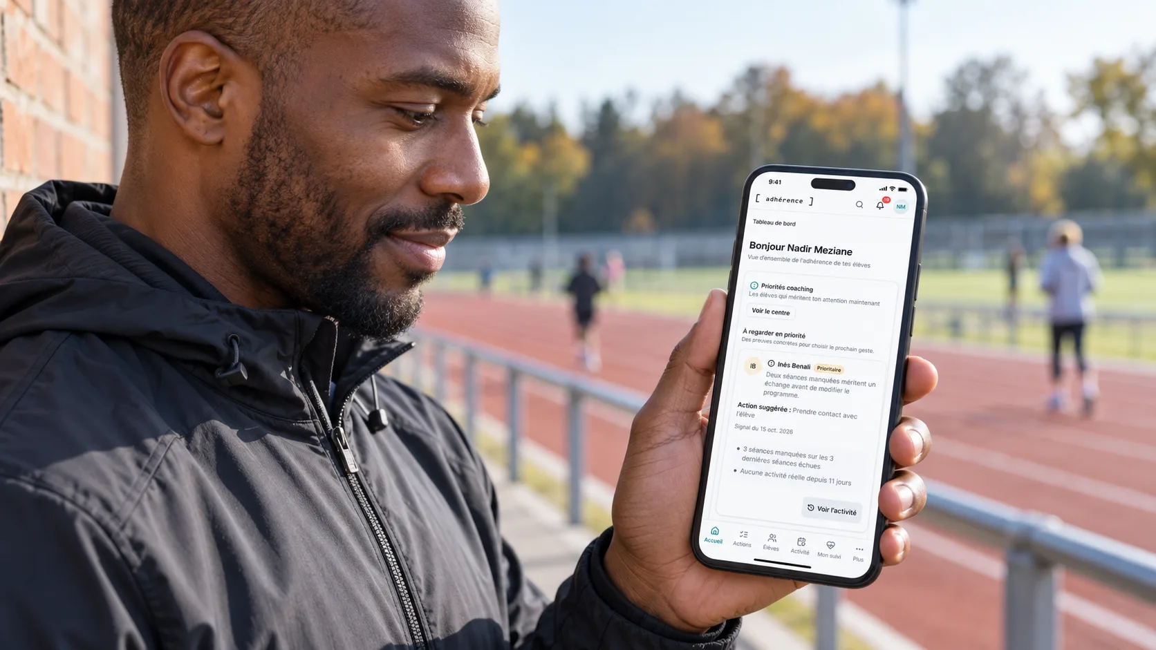 Trainer checking Adhérence coaching priorities on a phone beside an athletics track between appointments.