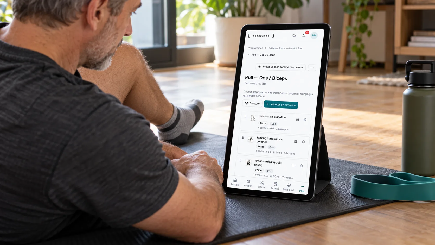 Coach relisant une liste d’exercices Adhérence sur tablette, assis sur un tapis dans un espace d’entraînement à domicile.