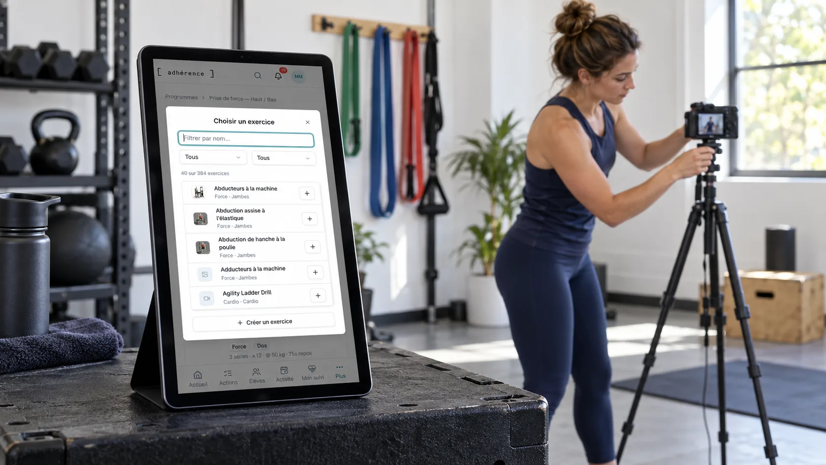 Catalogue d’exercices Adhérence sur une tablette au premier plan pendant qu’une coach prépare un tournage dans son studio.