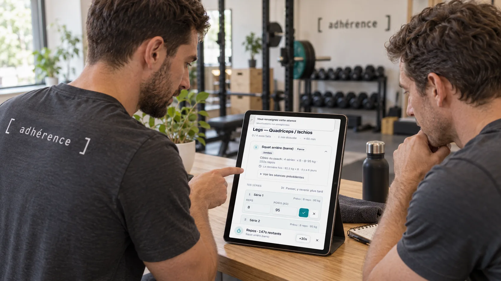 Coach et élève regardant les séries et charges d’une séance Adhérence sur une tablette en salle.