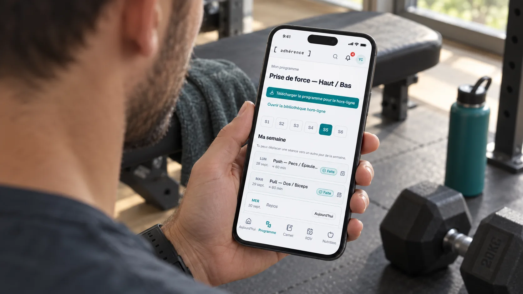 Élève consultant son programme Adhérence sur un téléphone près d’un banc de musculation.